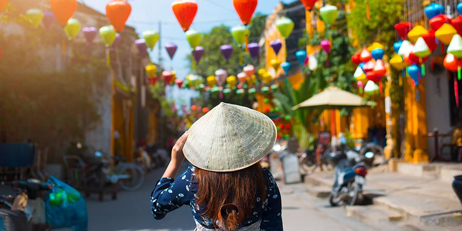 A woman wearing a traditional Vietnamese straw hat walks through a street decorated with colourful lanterns.