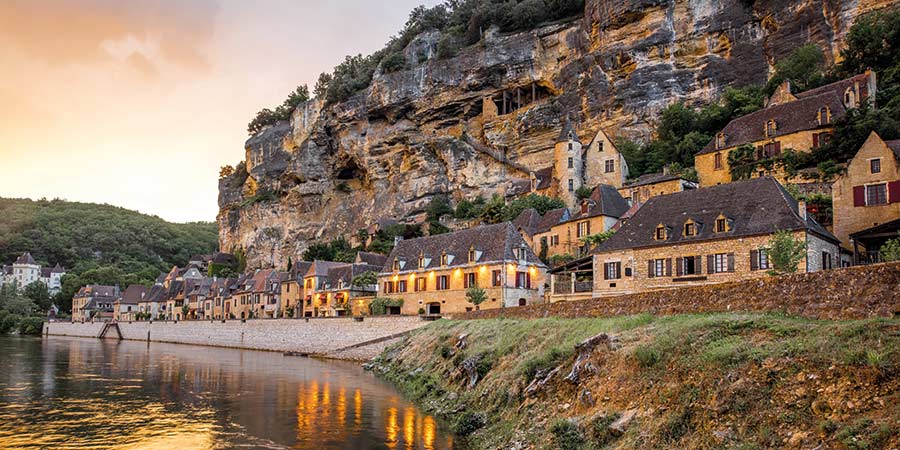 At the foot of a rocky cliffside, the picturesque commune of La Roque-Gageac is reflected in the water of the Dordogne river.